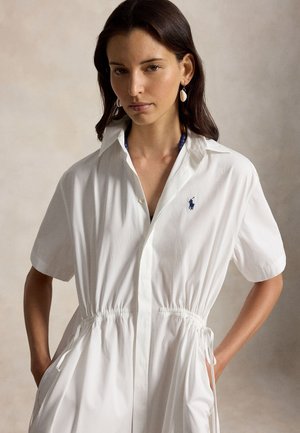 Kobieta o ciemnych włosach, ubrana w białą sukienkę z krótkim rękawem z sznurkiem w talii i kolczyki w kształcie muszli, pozuje z rękami w kieszeniach na neutralnym tle.