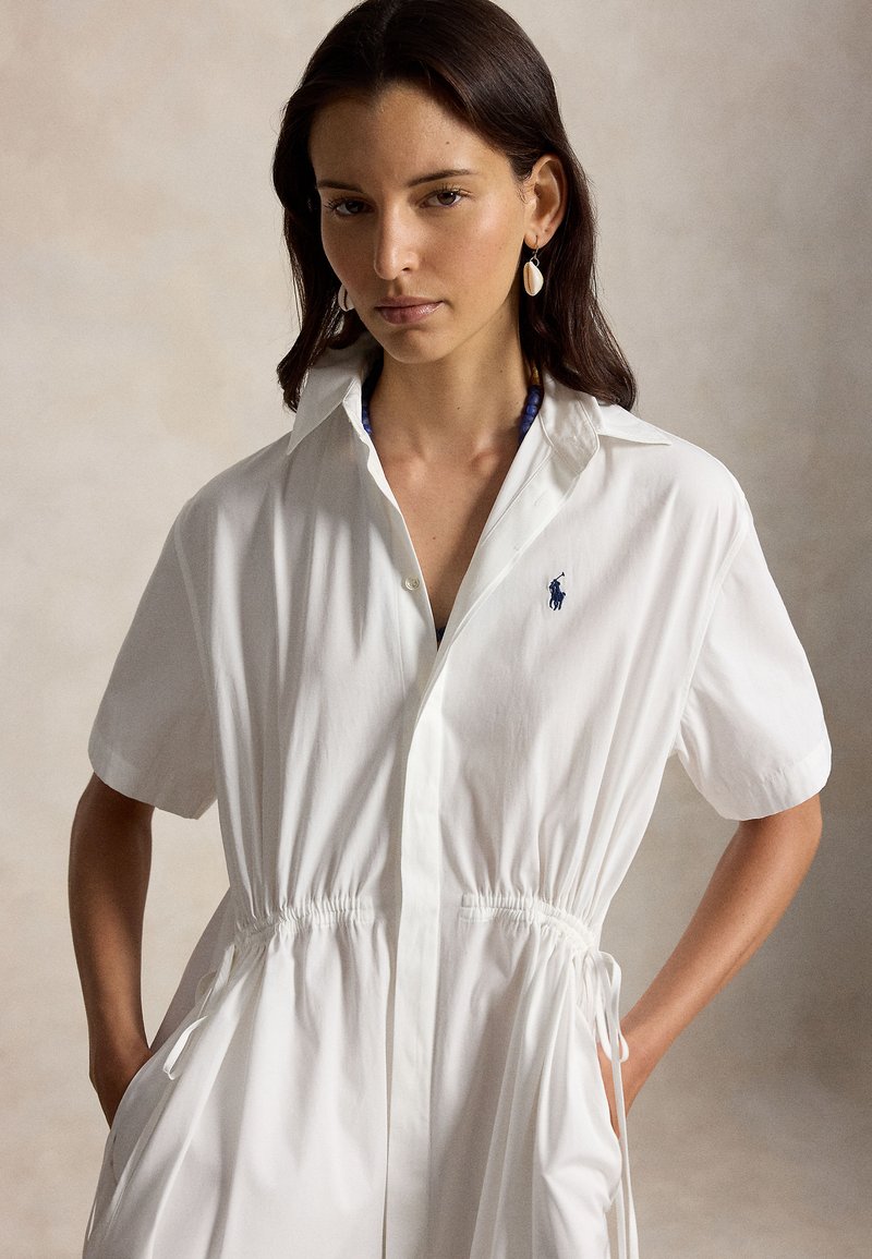 Femme aux cheveux foncés portant une robe blanche à manches courtes avec taille à cordon et boucles d'oreilles en forme de coquillage, posant les mains dans les poches devant un fond neutre.
