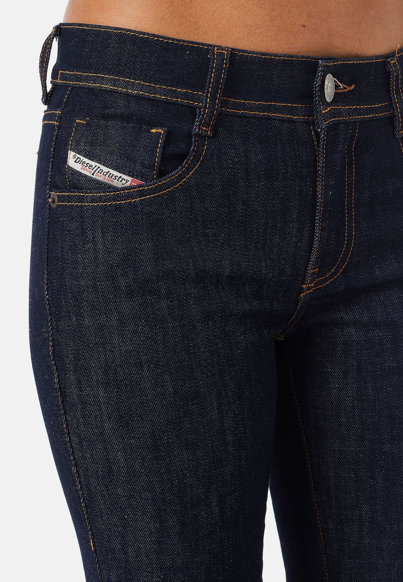 Vaqueros de mezclilla azul oscuro con un diseño ajustado. Las características incluyen costuras naranjas, un bolsillo frontal con una etiqueta de logo y un cierre clásico de botón.