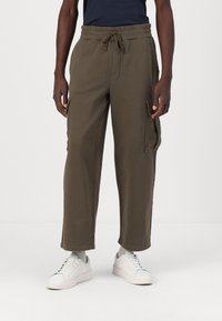Dark olive green cargo pants with elastic waist and drawstring, featuring side pockets and a relaxed fit. Paired with white sneakers.