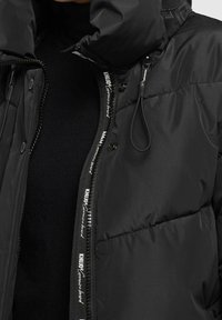 Černá puffer bunda s vysokým límcem a kapucí na stahování, se zipovým zapínáním a logovým páskem vpředu. Hladká, prošívaná textura.