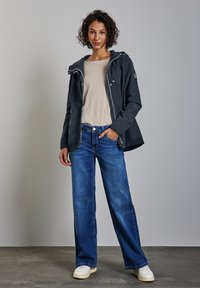 Svart hoodie-jacka med blixtlås, lätt tyg. Beige topp och vida blå jeans. Vita sneakers med ljus sula. Enkelt, praktiskt design.
