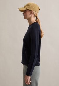 Marineblauer Pullover mit langen Ärmeln und rundem Ausschnitt, kombiniert mit hellgrauen Hosen. Ein beigefarbener Baseballcap und silberne Creolen runden den Look ab.