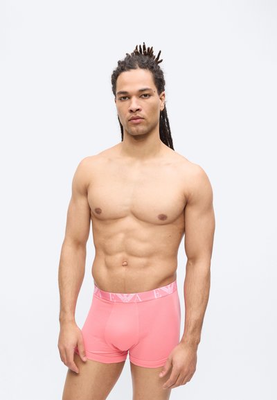 Μυώδης άνδρας με dreadlocks που φοράει ροζ αθλητικό εσώρουχο Armani, στέκεται μπροστά σε μια απλή φωτεινή επιφάνεια.