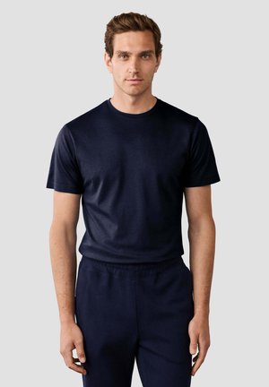Junger Mann mit kurzen braunen Haaren, der ein schlichtes dunkelblaues T-Shirt mit kurzen Ärmeln und passende Hosen trägt, steht vor einem einfarbigen Hintergrund.