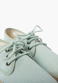 Scarpe di tela verde chiaro con punta rotonda, occhielli in metallo e lacci coordinati. Suola in fibra naturale con dettagli cuciti.
