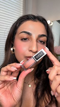 Lipgloss in een doorzichtig tube met een roze-nude tint. De verpakking heeft een zwart etiket met "IDOLÉ" in witte letters. Handen tonen lange, netjes onderhouden nagels.