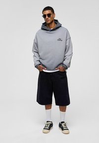 Grijze oversized hoodie met een zwarte "Stay Moment" print, gecombineerd met zwarte shorts, witte sokken en zwarte sneakers met crème accenten.