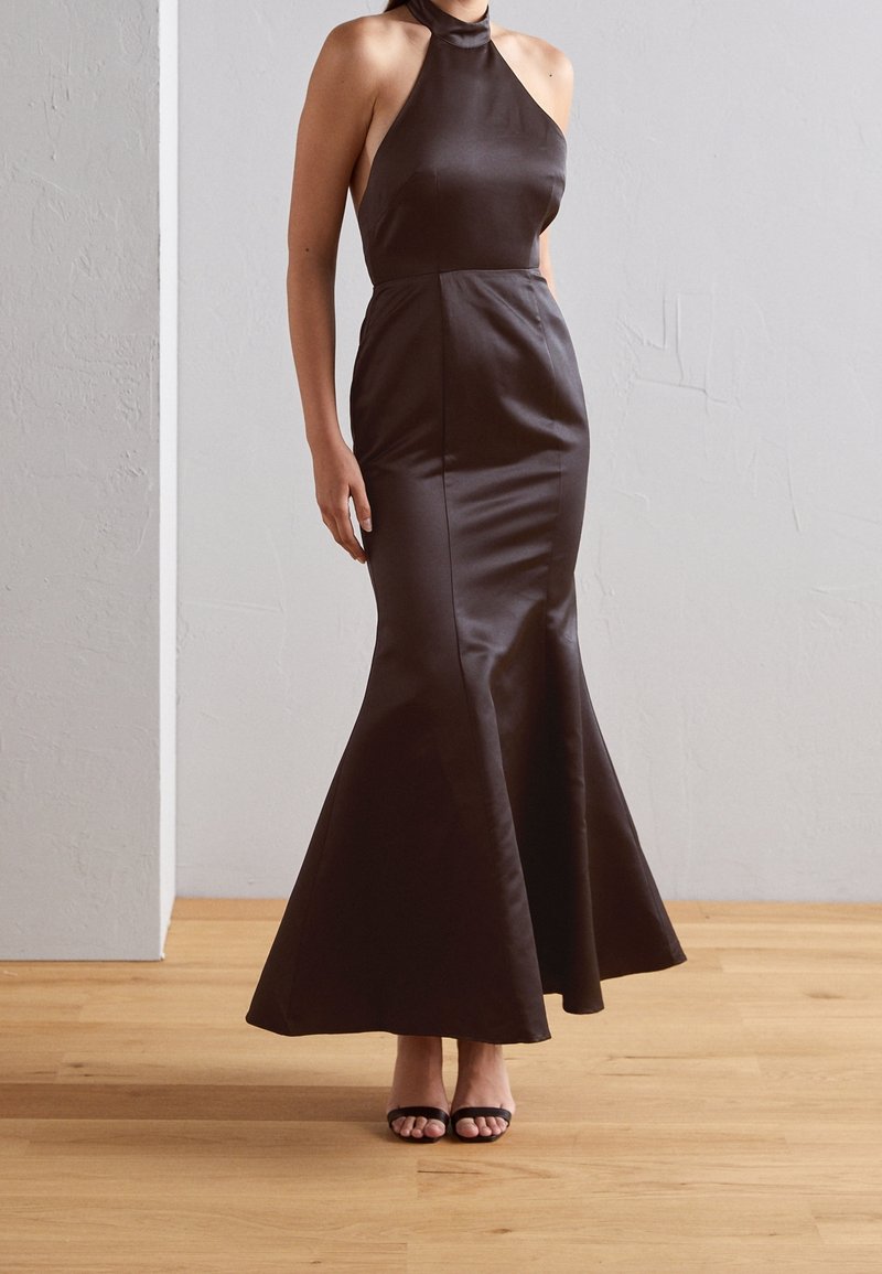 Femme portant une robe sirène en satin brun foncé sans manches avec un décolleté licou et des talons noirs à brides, debout sur un sol en bois.