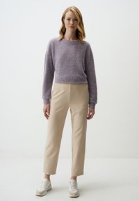 Maglione viola morbido con maniche lunghe, abbinato a pantaloni beige a vita alta. Il modello indossa sneakers bianche e beige. Sfondo minimalista.