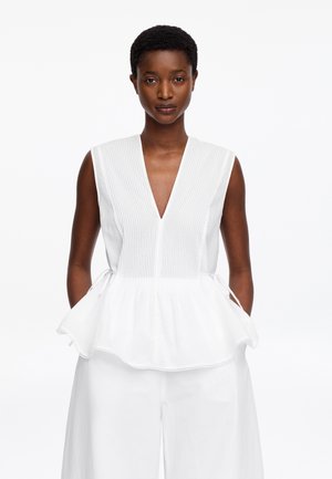 Femme portant un haut blanc sans manches avec un col en V plissé et une taille péplum, associée à un pantalon blanc à jambes larges, debout sur fond blanc.