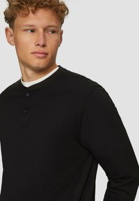 Schwarzes Langarm-Henley-Shirt mit Rundhalsausschnitt, dreiknöpfiger Knopfleiste und strukturiertem Stoff. Das Design ist schlicht und zeitgemäß.