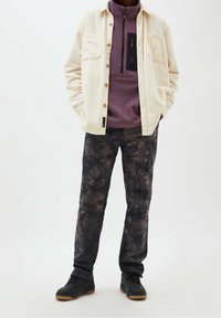Beige corduroy overhemd met knopen over een paarse fleece pullover met een borstzak, gecombineerd met zwarte tie-dye broek en zwarte schoenen.