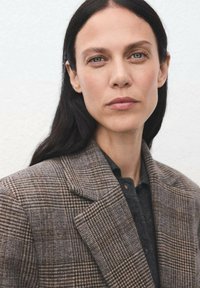 Brązowa marynarka w kratę z wyraźnie zarysowanymi ramionami, teksturowanym materiałem i klapami w kształcie notcha, noszona na ciemnej koszuli zapinanej na guziki.