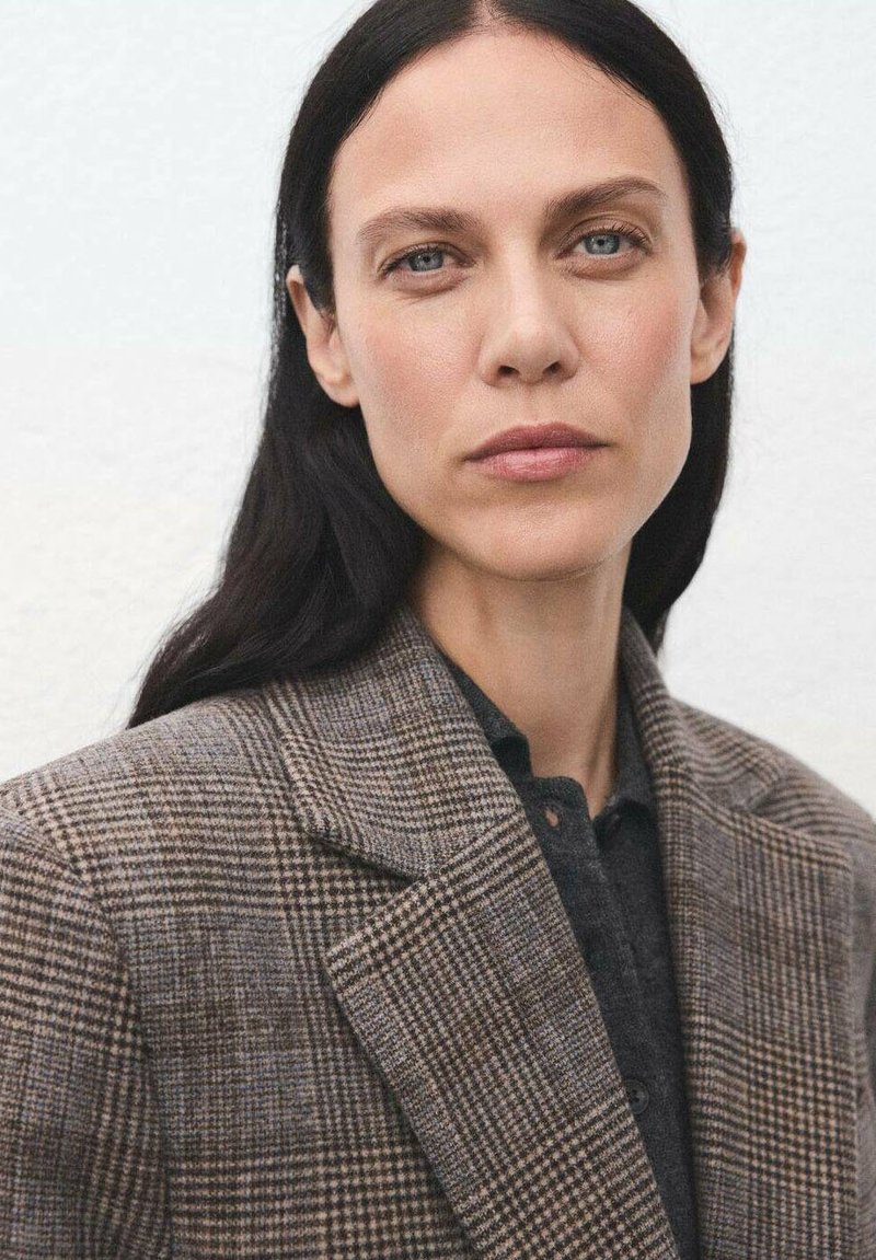 Brązowa marynarka w kratę z wyraźnie zarysowanymi ramionami, teksturowanym materiałem i klapami w kształcie notcha, noszona na ciemnej koszuli zapinanej na guziki.