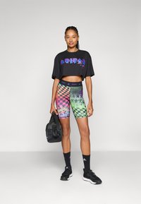 Černé zkrácené tričko s fialovým textem, barevné kostkované šortky na kolo v růžové, zelené a oranžové barvě, doplněné černými atlety a taškou.