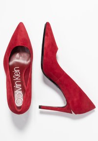 Pompes à talons hauts en suede rouge avec des bouts pointus, présentant un talon noir brillant et une semelle intérieure marquée. Texture lisse avec un design épuré.