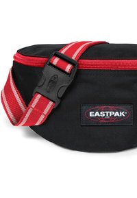 Czarna torba na biodro o okrągłym kształcie, z czerwonym paskiem w paski i czarną klamrą. Posiada naszywkę z logo Eastpak. Gładka tekstura, zapinana na zamek.