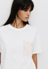 Witte katoenen t-shirt met een kort mouw ontwerp, voorzien van een gestructureerde beige kantzak aan de linkerkant.