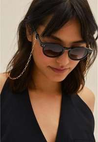 Černé sluneční brýle s tmavými skly, které mají jedinečný tvar a zlaté řetízkové uchycení. Elegantní design s lehkou texturou a minimálním zdobením.