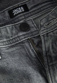 Grå denimtyg med en synlig dragkedja och knappstängning. Har en "JACK & JONES"-etikett. Texturen visar en slät yta och en subtil lyster.