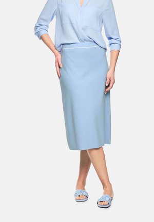 Frau trägt ein hellblaues Hemd mit Knöpfen, einen passenden knielangen Rock und offene Sandalen mit dekorativen Ausschnitten.