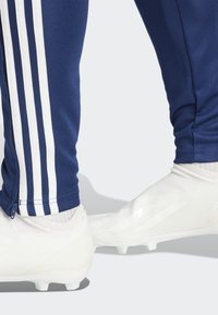 Námornícke športové nohavice s bielym trojprúžkovým dizajnom na stranách, spárované s bielymi štíhlymi futbalovými kopačkami s textúrovaným povrchom a výrazným brandingom.