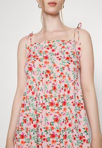 Vestido rosa com padrão floral, alças finas, decote franzido e detalhes plissados. Apresenta vibrantes designs de flores vermelhas e verdes.