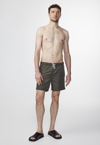 Shorts de bain vert foncé avec une taille élastique et un cordon de serrage, fabriqués en tissu lisse. Le modèle est pieds nus et porte des sandales noires.