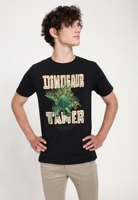 Mladý muž s kudrnatými vlasy, oblečený v černém tričku s obrazem zeleného stegosauruse a textem "Dinosaur Tamer", má ruce na bocích a dívá se doprava.
