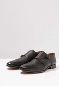 Svarta läderskor med en elegant design, som har dekorativa perforeringar och dubbla spännen på ovansidan. Rund tå och låg klack.