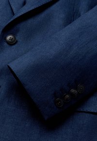 Marineblaue Blazerärmels mit vier schwarzen Knöpfen und Teil des Vorderteils, der einen Knopf mit der Aufschrift "Hackett London" zeigt.