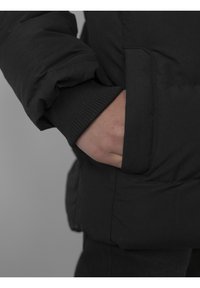 Doudoune noire avec un poignet côtelé, dotée d'une poche latérale où une main est posée. Le matériau semble lisse et texturé.