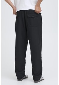 Pantaloni de trening negri cu croială lejeră, având un buzunar în spate cu închidere cu nasture, asortați cu șlapi cu dungi.