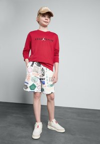 Rød langermet skjorte med "JORDAN" logo, mønstrede hvite shorts med ulike grafikker, beige caps, og hvite sneakers med detaljer.