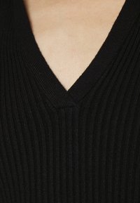 Indumento nero a coste con scollatura a V. La texture è prominente con leggere variazioni di spessore in tutto il capo.