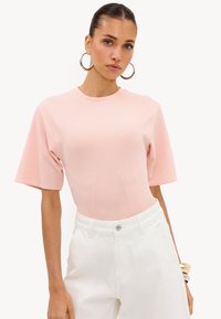 Femme aux cheveux plaqués en arrière portant de grandes boucles d'oreilles, un haut rose clair à manches courtes et un pantalon blanc taille haute, debout sur un fond uni.