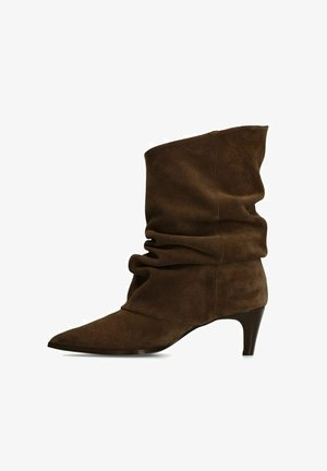 Botte molle en daim marron montant jusqu'au milieu du mollet, avec bout pointu et petit talon courbé, vue de profil sur fond blanc.