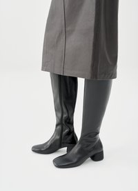 Juodi aukšti iki kelių odiniai batai iš lygiapluoštės odos. Kvadratinės noselės ir storas kulnas. Papildyti pilkos spalvos odine suknele su švariu apdaila.