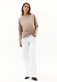 Beige Oberteil mit langen Ärmeln kombiniert mit weißen Schlagjeans. Das Outfit umfasst einen strukturierten Gürtel und schwarze Ballerinas. Schlichtes, modernes Design.