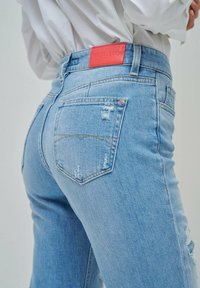 Jeans de mezclilla azul claro con un corte de altura media, detalles desgastados en las piernas y un parche de logo de cuero rojo en la cinturilla.