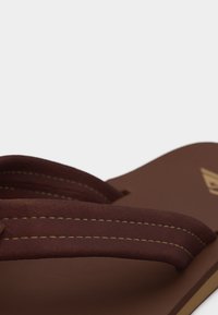 Καφέ σανδάλι τύπου φλιπ-φλοπ με διπλή ραφή στα λουράκια και μπεζ λογότυπο στο πέλμα, εμφανίζεται σε κοντινή λήψη με λευκό φόντο.