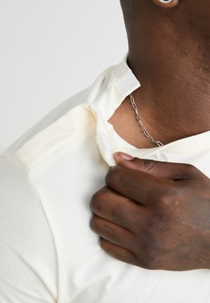 Cremefarbenes kurzärmliges Hemd mit einem Druckknopfverschluss am Hals und einer metallischen Kette, die unter dem Kragen sichtbar ist und Textur sowie Details zeigt.