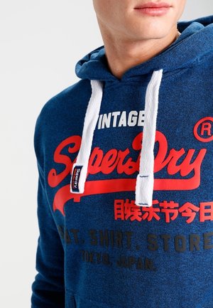 Mężczyzna w niebieskiej bluzie z kapturem SuperDry z białymi sznurkami i czerwono-biało-czarnym napisem, w tym "Vintage SuperDry" oraz japońskie znaki.