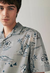 Rövid ujjú, világosszürke gombos ing sötétkék trópusi mintával, amely pálmafákat, vitorlás hajókat és "Aloha" feliratot ábrázol, puha anyagból készült.