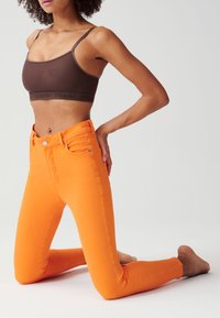 Hoge taille, aansluitende oranje jeans met gestikte details en voorkruiszakken, gecombineerd met een bruine sportbeha met een logo band.