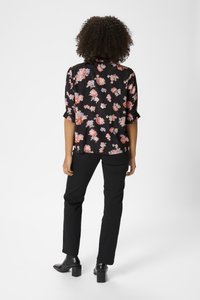 Blouse noire à motifs floraux roses et violets, manches courtes retroussées, coupe décontractée, associée à un pantalon noir et des bottes à talons épais.