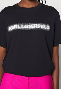 Camiseta de algodón negra con el texto "KARL LAGERFELD" en puntos blancos en el pecho. Mangas cortas y cuello redondo. Textura suave.