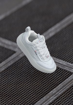 Hvit sneaker med en glatt overflate, teksturert sidesdesign og tykk såle. Har snøring og en logo på tungen for merkeidentifikasjon.