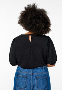 Blusa nera con texture, maniche corte a palloncino e apertura a occhiello sul retro, indossata con jeans in denim blu a vita alta.